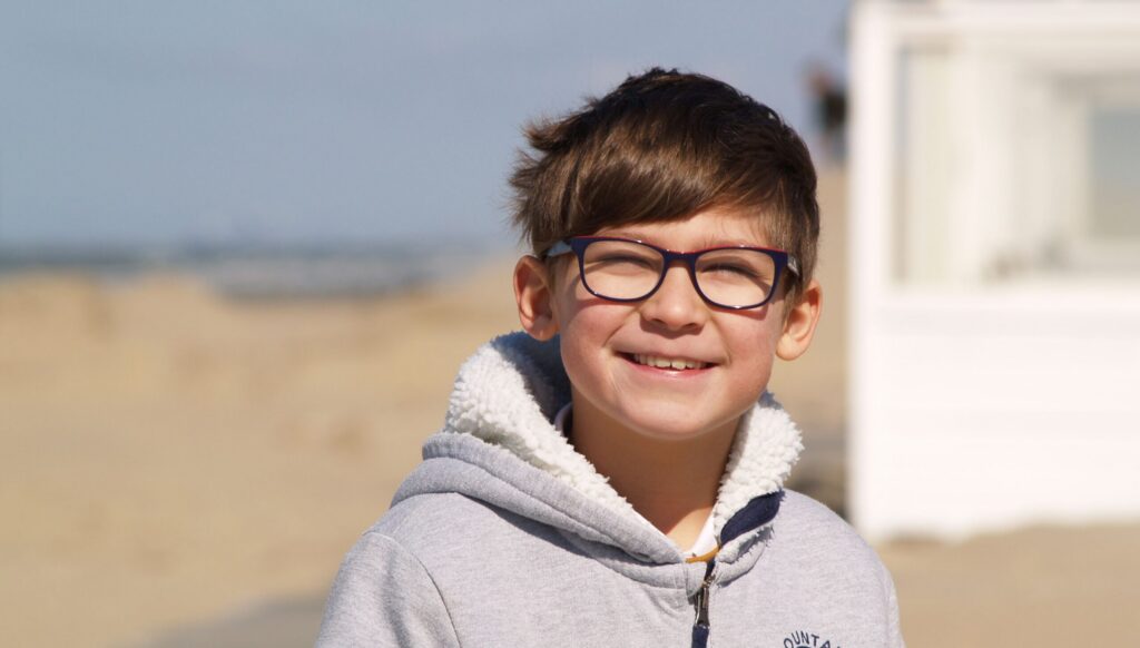 A cheerful child with glasses smiles at the beach, wearing a cozy hoodie on a sunny day.